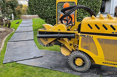 Forestry machinery driving on black HDPE ground protection mats laid over a green lawn to prevent turf damage.