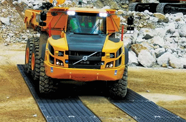Heavy mining dump truck driving on black HDPE ground protection mats, creating a stable temporary haul road at a rocky mining site.