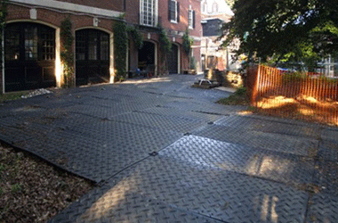 Black interlocking HDPE ground protection mats with anti-slip diamond tread laid over a yard at a residential construction site.