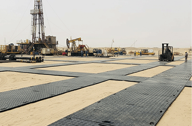 Extensive network of black HDPE ground protection mats laid on sand, creating a stable working platform for an oil and gas drilling rig.