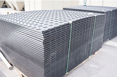 Large stacks of black HDPE ground protection mats with anti-slip diamond tread, neatly organized in a factory for bulk shipment to the rental industry customers.